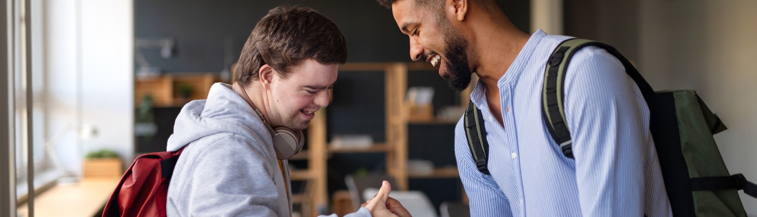 Young happy man with Down syndrome with his mentoring friend greeting indoors at school.